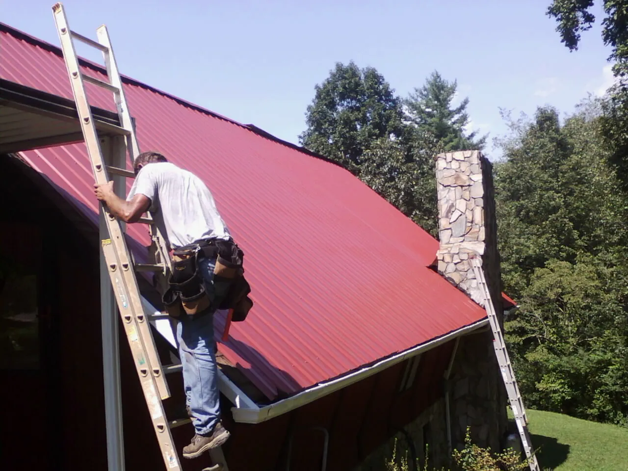 Roofer on a ladder beside a red metal roof and stone chimney — Soffit & Fascia Repair by Roofing Brothers in Indiana